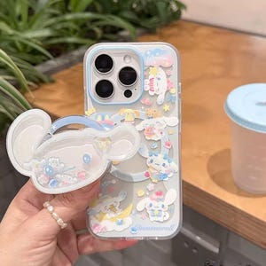 May include: Clear phone case with a cartoon design featuring a white character with blue accents. The case has a matching pop-up grip. The design includes the word "Dream" and the word "Cinnamoroll". A light blue and pink drink cup is in the background.