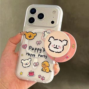 Könnte beinhalten: Transparente Handyhülle mit einem Cartoon-Welpen-Design. Die Hülle zeigt den Text "Puppy's Happy Party" und enthält Illustrationen von Welpen, einem Kuchen und Pfotenabdrücken. Ein passender rosa Pop-Socket mit einem Welpengesicht ist angebracht.