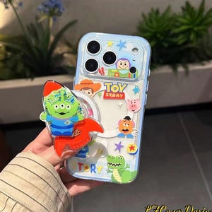 Puede incluir: Funda transparente para teléfono con borde azul, decorada con personajes de "Toy Story". La funda presenta imágenes de Buzz Lightyear, Woody y otros personajes. Un extraterrestre verde en un cohete está adherido a la funda.