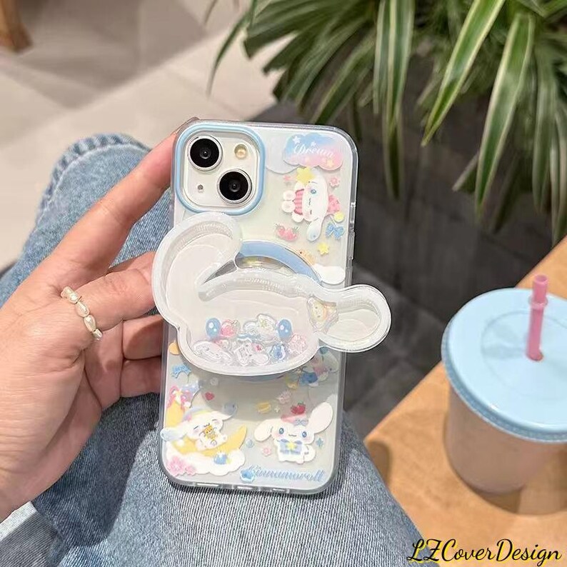 May include: A clear phone case with a blue border and a white, cartoon-style character design. The case features a pop-up grip and is decorated with small, colorful embellishments. The text "Cinnamoroll" is visible on the case.