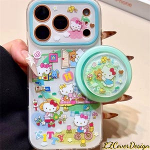 以下が含まれることがあります： ライトブルーの縁取りとポップアップグリップが付いた透明な携帯電話ケース。ケースは、ハローキティやその他の漫画のキャラクターで庭のシーンが描かれています。ポップアップグリップはキラキラしたデザインです。