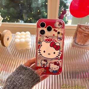 May include: A red and clear phone case featuring Hello Kitty designs. The case has a red border and a clear back, showcasing various Hello Kitty illustrations with red bows. The case is designed for a phone with multiple camera lenses.