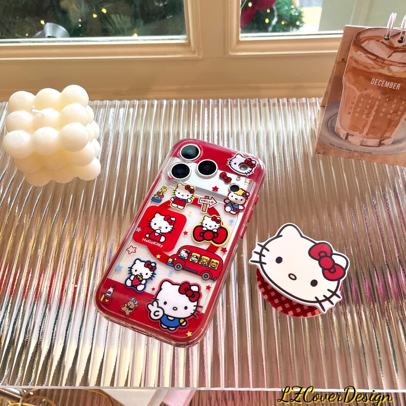 May include: A clear phone case with a red border featuring Hello Kitty designs. The case has multiple images of Hello Kitty, a red bus, and stars. A Hello Kitty phone grip and a white candle are also visible.