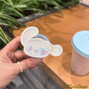 May include: A clear, bunny-shaped hair clip filled with small, colorful charms. The clip is held in a hand, with a light blue cup with a lid in the background. The clip has a blue handle.