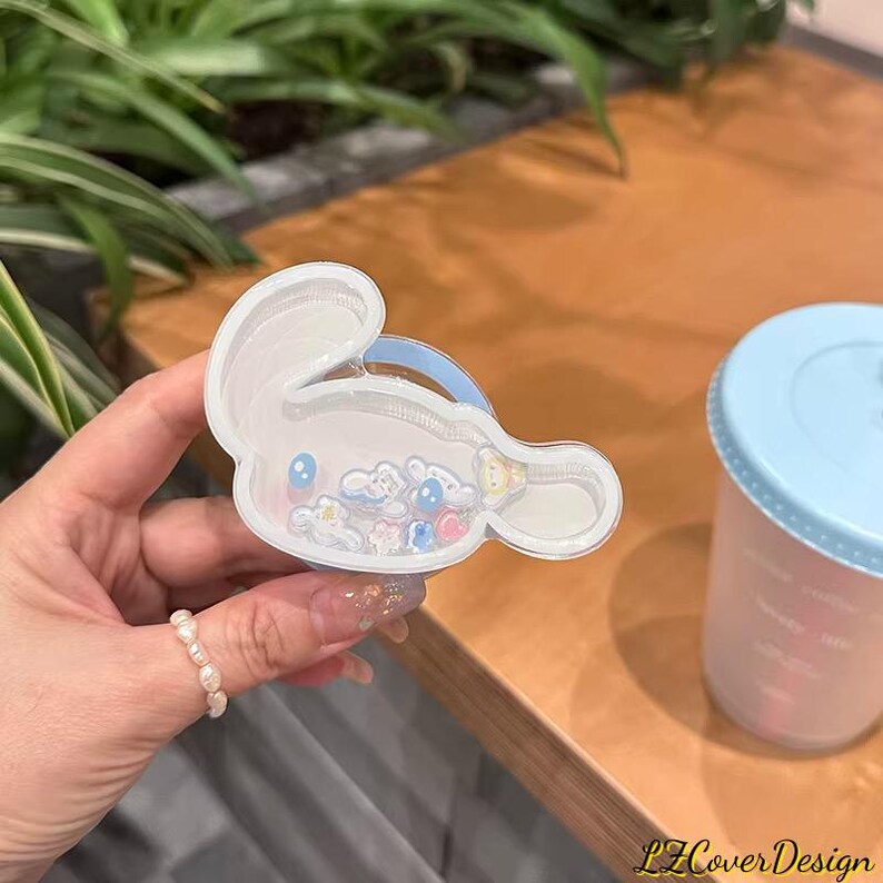 May include: A clear, bunny-shaped hair clip filled with small, colorful charms. The clip is held in a hand, with a light blue cup with a lid in the background. The clip has a blue handle.