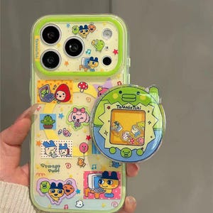 May include: A clear phone case with a lime green border and a colorful design featuring cartoon characters and the text "Tamagotchi". A matching Tamagotchi-themed phone grip is attached.