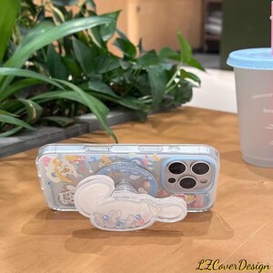 May include: A clear phone case with a Cinnamoroll design, featuring a matching pop socket. The case has a light blue border and is decorated with cartoon characters. The pop socket is white and shaped like Cinnamoroll.