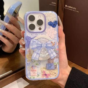 May include: A decorative phone case with a light blue and purple color scheme. The case features a bow, floral accents, and a pop-up grip. The design includes a collage of images and text, with a butterfly and blueberries. The case is designed for a phone.