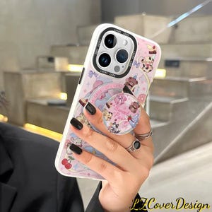 May include: A pink and white phone case with a pop socket featuring a cartoon design of bears, cherries, and donuts. The case has the text "LUCKY CASE" and "Sweet Party". The phone case is held by a person.