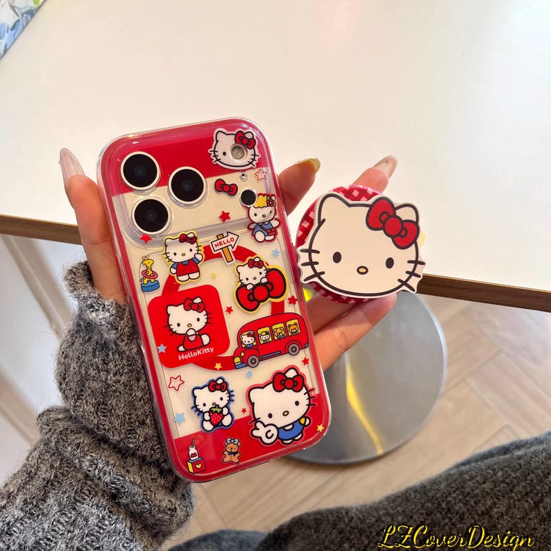 May include: A red and clear phone case featuring Hello Kitty designs and a matching pop socket. The case has a red border and showcases various Hello Kitty illustrations, including the character on a bus and holding a strawberry. The pop socket has a white background with Hello Kitty's face.
