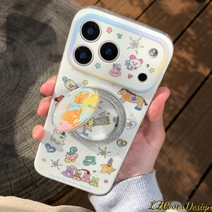May include: A white phone case with a whimsical design featuring cartoon animals, stars, and other playful elements. The case has a built-in pop socket with a glittery silver ring and a colorful bunny illustration. The case is designed for a smartphone.