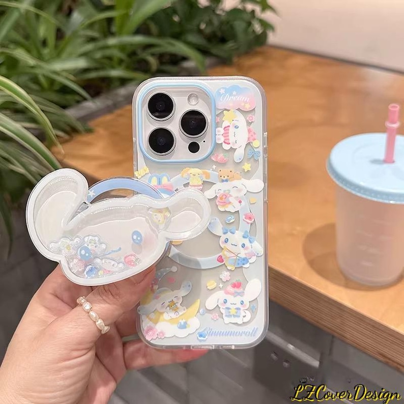 May include: Clear phone case with a cartoon design featuring a white character with blue accents. The case has a matching pop-up grip. The design includes the word "Dream" and the word "Cinnamoroll". A light blue and pink drink cup is in the background.