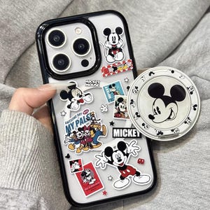 May include: Clear phone case with black edging, decorated with Mickey Mouse stickers and a matching pop socket. The stickers show different Mickey Mouse poses and phrases. The pop socket has a white background with a Mickey Mouse face.