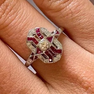 Könnte beinhalten: Ein silberfarbener Ring mit einem zentralen runden weißen Diamanten, umgeben von geometrischen Mustern aus tiefroten Rubinen und kleinen, im Pavé-Stil gefassten weißen Diamanten. Die Metallschiene ist schmal und poliert und zeigt ein komplexes, längliches Design.