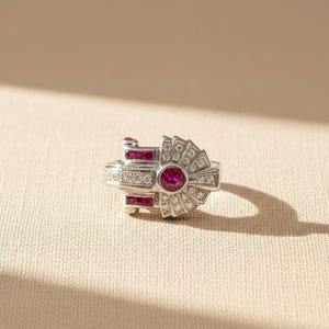 May include: A silver ring featuring a central round-cut ruby surrounded by small white diamonds. The design includes a fan-shaped section set with diamonds and two rectangular segments containing square-cut rubies on the sides of the band.