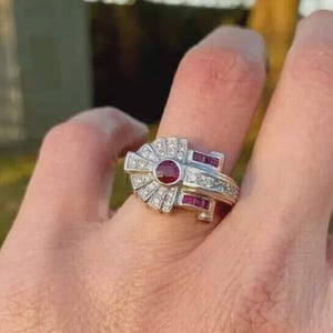 Könnte beinhalten: Ein silberfarbener Ring mit einem zentralen runden roten Edelstein, der von einer fächerförmigen Anordnung kleiner klarer Steine umgeben ist. Die Ringschiene enthält zusätzliche rechteckige rote Steine, die seitlich in das Metall eingelassen sind.