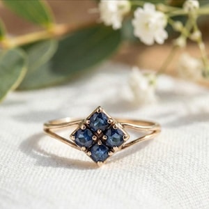 May include: A gold ring featuring a cluster of four square-cut dark blue sapphires arranged in a diamond shape. The band is thin and splits into two delicate strands as it reaches the central gemstone setting, creating a refined and balanced look.