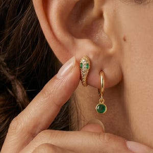 Può includere: Primo piano di un orecchio adornato da tre orecchini d'oro. Uno è un piccolo cerchio con una pietra verde, un altro è un orecchino a forma di serpente con pietre verdi e trasparenti, e il terzo è un gruppo di pietre verdi.