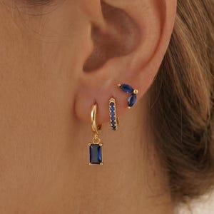 May include: Gold-toned earrings with blue gemstones. The earrings include a rectangular drop earring, a small hoop with blue stones, and a stud with a curved design of blue stones. The earrings are displayed on an ear.