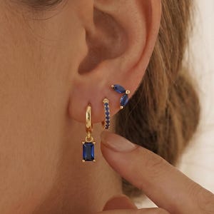 May include: A collection of gold earrings with blue gemstones. The earrings include a pair of small hoops with blue stones, a pair of stud earrings with three blue stones, and a pair of gold hoops with a rectangular blue gemstone pendant.