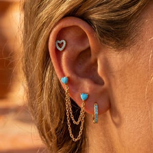 May include: A gold-toned ear piercing set featuring a small heart-shaped stud with clear stones in the upper cartilage, and two heart-shaped turquoise studs in the lobe connected by two delicate gold chains, plus a small gold hoop with clear stones.