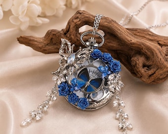 May include: A silver-toned pocket watch pendant on a chain, featuring a blue butterfly graphic on the watch face. The exterior is decorated with a silver butterfly, clusters of small blue roses, and dangling clear crystal leaf-shaped charms.