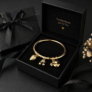 May include: A gold-colored bangle bracelet with three charms: a pineapple, cherries, and a heart. The bracelet is displayed in an open black gift box with the text "Timeless Beauty Just for You". A second black gift box with a bow is visible.