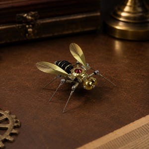 Peut inclure: Une figurine d'insecte mécanique construite à partir de composants métalliques. Le corps présente un abdomen noir segmenté, un thorax doré avec un accent rouge central et deux grandes ailes translucides en éventail. Il a des pattes fines.
