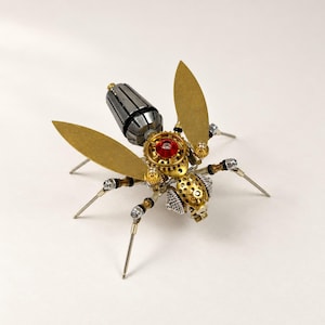May include: A mechanical insect sculpture constructed from metal components including gears, screws, and nuts. The body features a silver-toned segmented abdomen, a gold-toned thorax with a central red accent, and two large, flat, gold-toned wings.