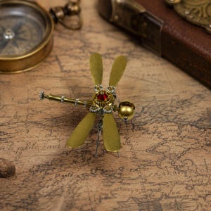 Może przedstawiać: Broszka w kształcie ważki wykonana z metalowych elementów w kolorze mosiądzu, posiadająca cztery płaskie skrzydła, segmentowy ogon oraz korpus ozdobiony małymi akcentami przypominającymi koła zębate i okrągłym czerwonym kamieniem.