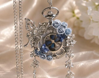 May include: A silver-toned pocket watch pendant on a chain, featuring a butterfly motif, clusters of light blue roses, and dangling clear crystal accents. The watch face is visible behind a decorative frame with Roman numerals and dark blue hands.