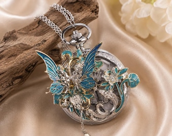May include: A silver-toned pocket watch pendant featuring an intricate overlay of blue and teal enamel butterfly and floral motifs. The design includes clear crystal accents, a delicate silver chain, and a small dangling charm, creating a detailed piece.