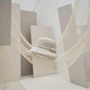May include: A beige and gray plaid cloche hat with a decorative bow, resting in a cream-colored net hammock. The hat has a rounded crown and a small brim. The background features neutral-toned geometric shapes.