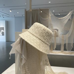 May include: Cream-colored bucket hat with a geometric pattern. The hat has a wide brim and is displayed with a sheer, draped fabric and a net-like material. The background features art and hanging fabric.
