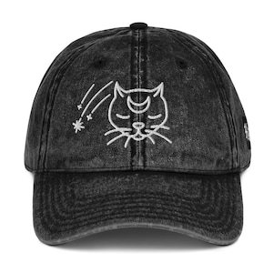 Könnte beinhalten: Schwarze Baseballkappe aus Denim mit einem weißen, gestickten Katzendesign, einem Mond und Sternschnuppen. Die Kappe hat einen gebogenen Schirm und einen verstellbaren Riemen. Ein modisches Accessoire.