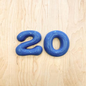 May include: A glossy, deep blue ceramic number 20. The number two and the number zero are shaped with rounded, smooth edges and a reflective surface finish, creating a bold and distinct visual representation of the numerical value twenty.