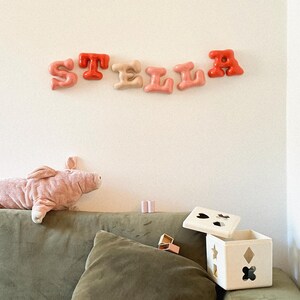 Op de afbeelding: Wandletters die het woord STELLA vormen in een glanzend, afgerond, bubbelachtig lettertype. De letters zijn gekleurd in tinten oranje, lichtbeige en roze. Daaronder een roze pluche varken en een witte houten vormensorteerdoos.