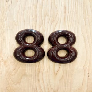 May include: Two identical dark brown, glossy, rounded number eight digits. Each digit features a smooth, reflective surface with a continuous, looped shape and a hollow center, creating a bold, three-dimensional appearance for display purposes.