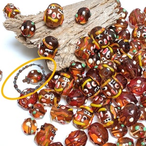 May include: A collection of brown glass beads featuring multi-colored abstract patterns, including white, yellow, and green dots and swirls. One bead is encircled by a thin, dark, beaded wire loop, highlighting its round shape and design.