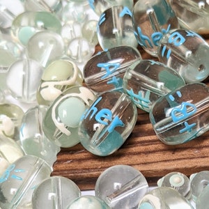 May include: A collection of clear glass beads with blue and white lettering. The beads are oval shaped and have a variety of different words written on them.