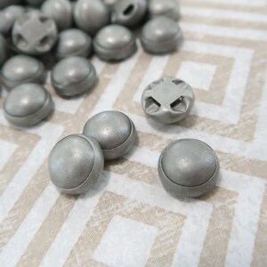 May include: A close-up of a group of silver buttons. The buttons are round and have a smooth, shiny finish. Some of the buttons have a decorative design on the top.