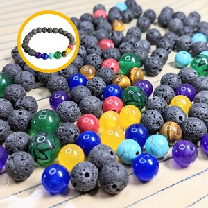 May include: A close-up of a variety of beads, including black lava rock beads, colorful gemstone beads, and tiger's eye beads. The beads are arranged in a random pattern on a white surface.