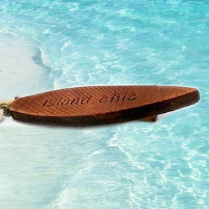 May include: A wooden surfboard charm with the words "island chic" engraved on it. The charm is brown and has a natural wood grain. It is strung on a beaded necklace with brown and white beads.