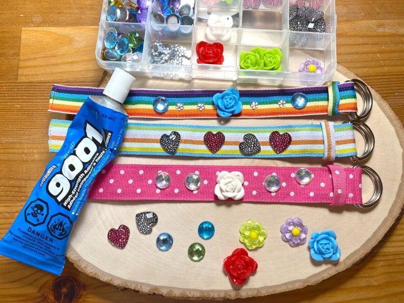 Puede incluir: Tres cinturones de cinta de colores con diferentes adornos, incluyendo corazones, flores y pedrer&iacute;a. Tambi&eacute;n se puede ver un tubo de pegamento en la imagen.