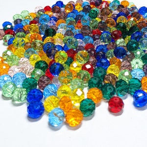 Puede incluir: Un gran surtido de cuentas de cristal facetadas de colores en varios tonos de azul, verde, rojo, amarillo y morado. Las cuentas están esparcidas sobre una superficie blanca.