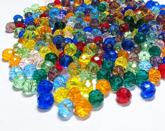 May include: A large assortment of colorful faceted glass beads in various shades of blue, green, red, yellow, and purple. The beads are scattered on a white surface.