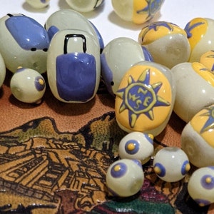 May include: A collection of small, oval-shaped glass beads with various designs. Some beads are white with blue and yellow accents, while others have a blue suitcase or a compass rose design. The beads are arranged on a brown surface with a pattern of a city.