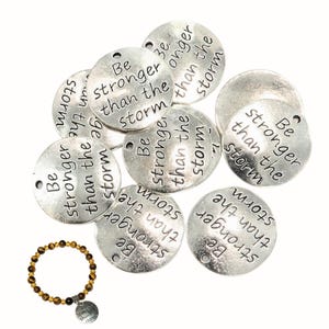 Puede incluir: Un conjunto de dijes redondos de metal plateado, cada uno grabado con el texto 'Be stronger than the storm' y con un pequeño orificio en la parte superior. También se incluye una pulsera hecha de cuentas de piedra ojo de tigre marrón y dorado.