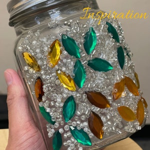 Puede incluir: Un tarro de vidrio decorado con hojas de vidrio verde, dorado y marr&oacute;n y cuentas blancas. El tarro tiene la palabra "Inspiration" escrita en el lateral.