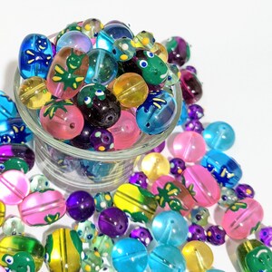 May include: A clear glass bowl overflowing with colorful glass beads. The beads are various shapes and sizes, including round, oval, and oblong. The beads are in shades of blue, green, pink, purple, and yellow, some with painted designs.